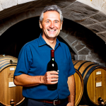 An experienced male winemaker, mid-50s, with a warm, genuine smile. He is dressed in a modest, professional work shirt and dark trousers, standing in a historic Italian wine cellar. The cellar is characterized by vaulted stone ceilings and numerous large, aged oak barrels that stretch into the soft, ambient light. He is holding a single wine bottle, gesturing gently as if explaining its story. The atmosphere is rich with tradition and quiet reverence. Perfect anatomy, correct proportions, natural pose, well-formed hands, proper finger count, natural body proportions. Professional photography, high quality, authentic, fully clothed, modest clothing, appropriate attire, professional dress, safe for work, appropriate content, family-friendly.